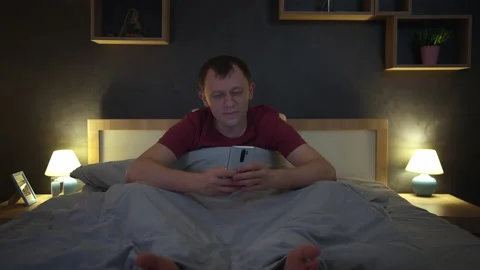 Man sitting in bed at night uses his mobile phone, browses the app, bedroom Stock Footage 216701199