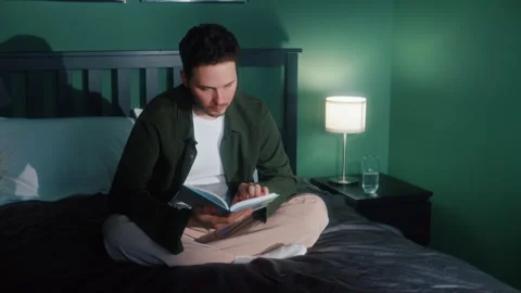 Man sitting on bed reading book in cozy bedroom Vídeos de archivo 319544600