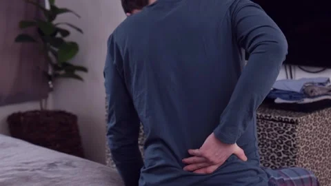 A man sitting on a bed rubs or massages his back for pain 库存影片 330474080