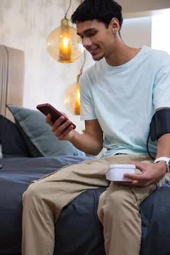 Man sitting on bed in softly lit bedroom holding smartphone, white monitor and Stock Photos