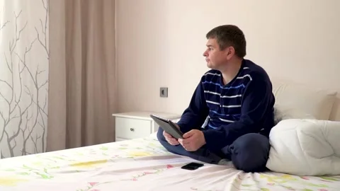 A man is sitting on the bed with a tablet and a phone. Real people, hommade Stock Footage 132040721
