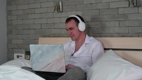 A man is sitting on the bed, using a laptop and wearing headphones. Man Stock Footage 278103964