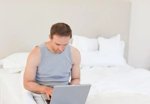 Man sitting on bed using silver laptop in neutral bedroom, wearing gray 写真素材