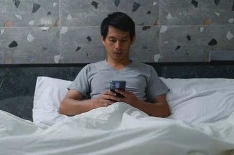 Man Sitting on Bed Using Smartphone in Cozy Bedroom Setting Photos