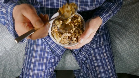 Man sitting on bed wearing pyjamas / pajamas eating a bowl of muesli. Video stock 129194971