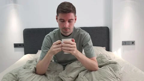 Man sitting on a bed wrapped in a warm blanket and drinking a hot beverage Video stock 284044780