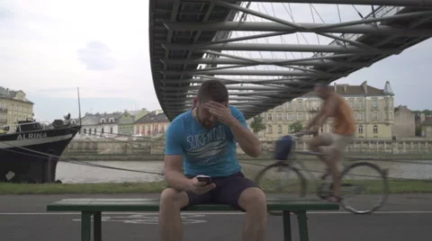 Man sitting on the bench against the river, receives bad message and walk away. 库存影片 65187778