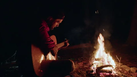 Man sitting on a bench and playing guitar in front of a campfire at night Stock Footage 266734319