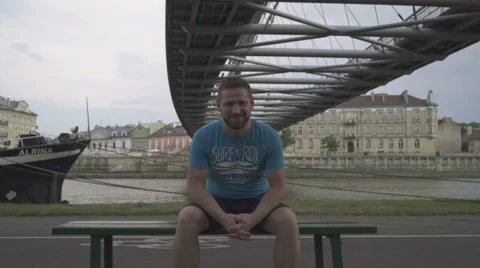Man sitting on the bench and smiling to camera against the river. Vídeo Stock 65187642