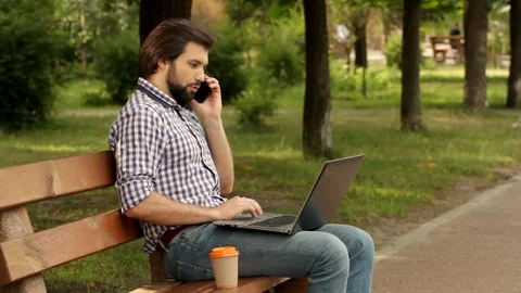 Man is sitting on bench and talking on phone. He is holding laptop on legs. Man Stock Footage 99740083