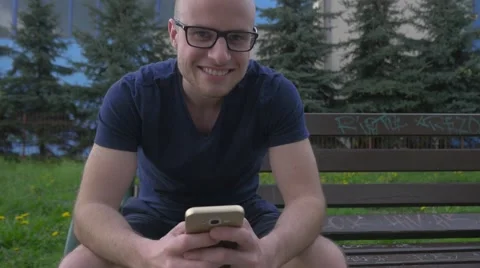 Man sitting on the bench and using phone. He looks into the camera and smile. Stock Footage 62520599