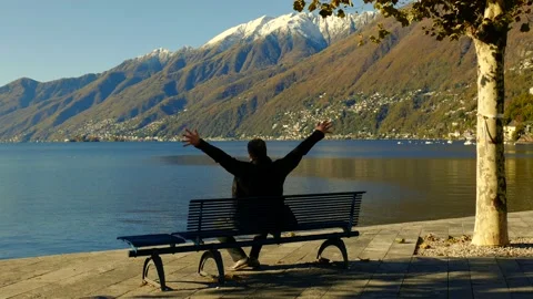 Man Sitting on a Bench with Arms Outstretched in front of Alpine Lake Stock Footage 164712982