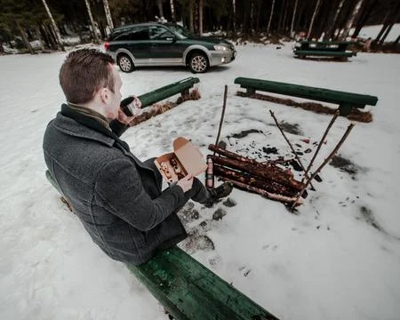Man Sitting on Bench by Fire Pit in Snowy Forested Area with SUV Nearby Stock Photos