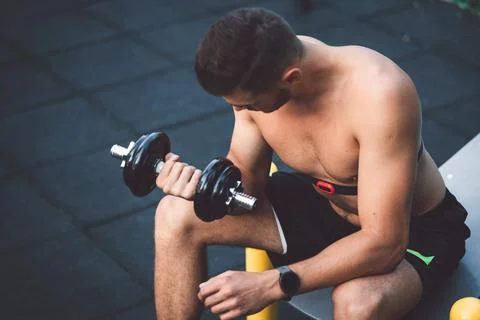 Man sitting on a bench lifting weights outdoors, while wearing a sports watch to Stock Photos