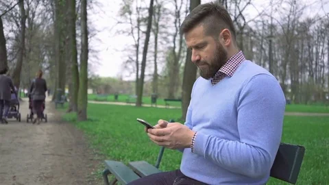 Man sitting on bench in park typing sms message by smartphone app, steadicam Video stock 74344130