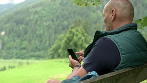 Man sitting on a bench in a scenic mountain landscape, using his smartphone Stock Footage 281045192