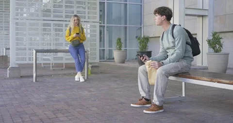 Man sitting on bench using phone, eating from paper bag, noticing woman in Stock Footage 340972411