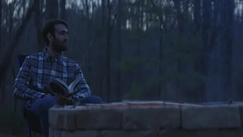 Man sitting beside a fire reading a book at dusk, enjoying nature Stock Footage 261438306