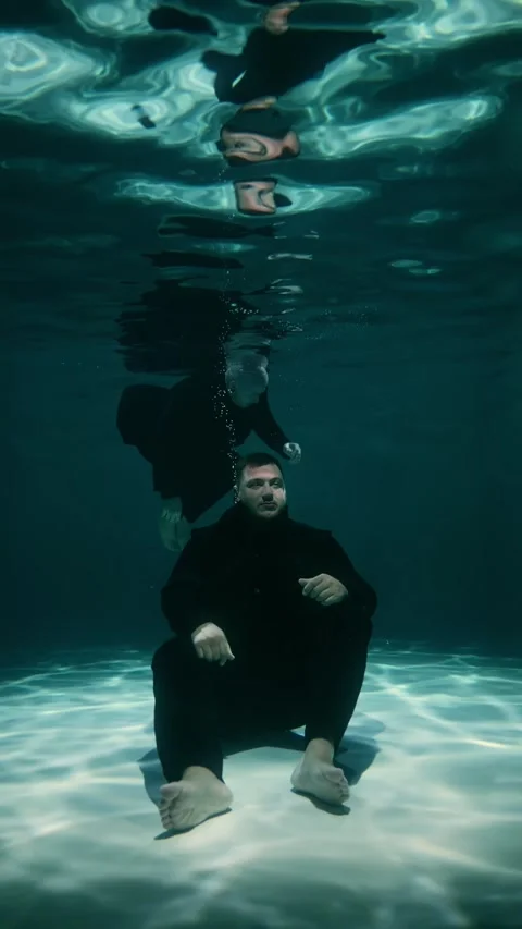 A man is sitting at the bottom of a pool underwater, another man unconscious Stock Footage 315559664
