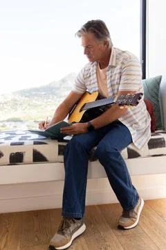 Man sitting on built-in window bench with acoustic guitar across lap, writing Stock Photos