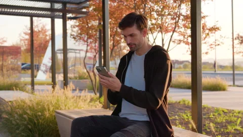 Man sitting at bus stop, checking smartphone, wearing casual attire, surrounded Stock Footage 315532449