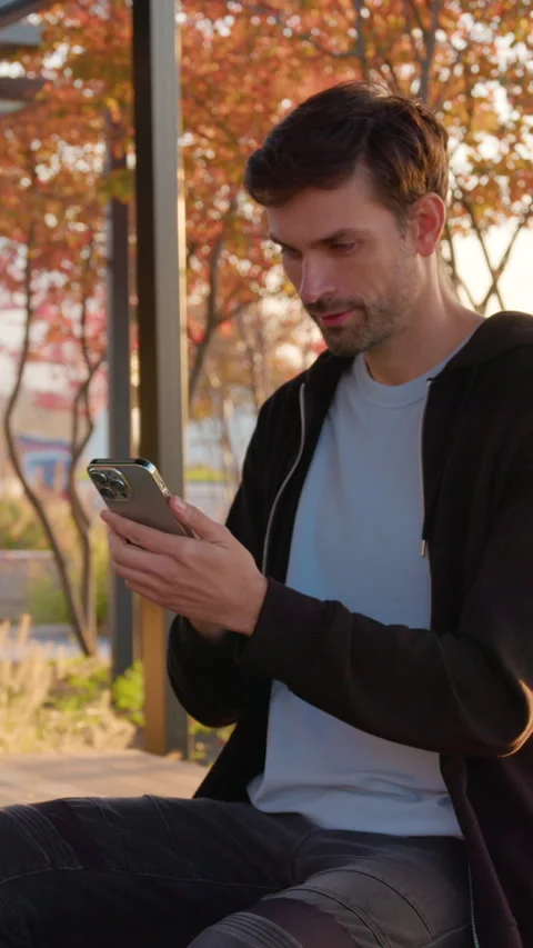 Man sitting at bus stop, checking smartphone, wearing casual attire, surrounded Stock Footage 318475702