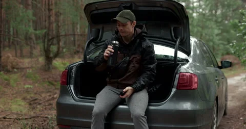 Man sitting on car trunk and drinking coffee from thermos with audio included Stock Footage 340845129
