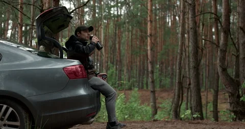Man sitting on car trunk and drinking coffee in pine forest, audio included Stock Footage 340845997