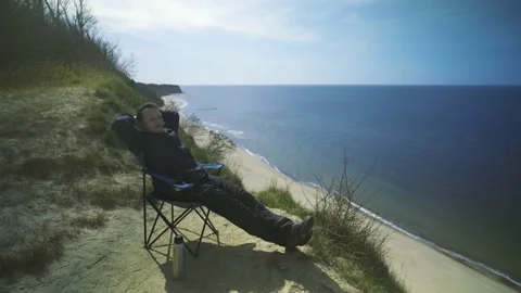 Man sitting in a chair drinking tea on a cliff by the sea. Stock Footage 239871275