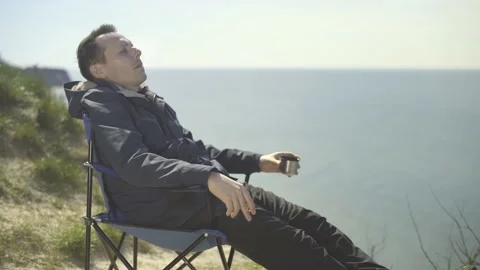 Man sitting in a chair drinking tea and smoking on a cliff by the sea. Stock Footage 239871541
