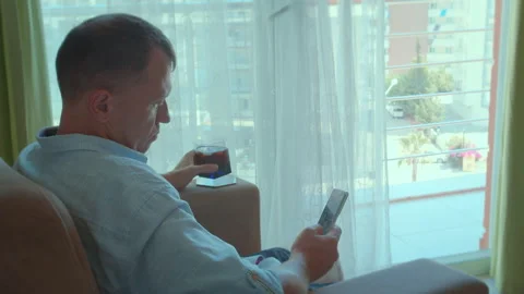 Man sitting in a chair in front of a window, using a smartphone app and drinkin Stock Footage 263999701