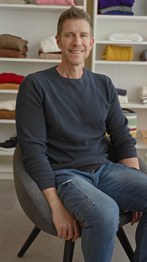 Man sitting on chair in studio with folded sweaters on shelves, smiling and.. Stock Footage 337389127