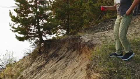 A man is sitting on a cliff and drinking a drink from a red mug Video stock 177993509