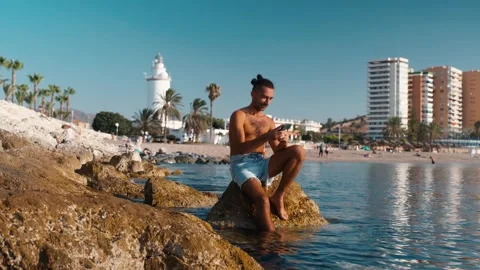 Man sitting on coastal rocks, engrossed in his smartphone, with a serene be.. Stock Footage 344059249