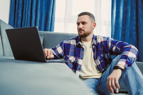A man is sitting on a couch and focused on a laptop screen while using a comp Stock Photos