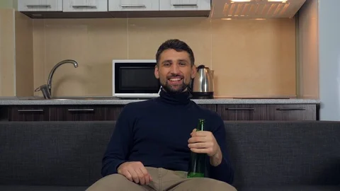 Man sitting on the couch, drinking beer, smiles and watches entertainment on TV Stock Footage 121840473