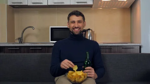 Man sitting on the couch, eats chips, drinking beer, smiles and watches TV Stock Footage 121841342