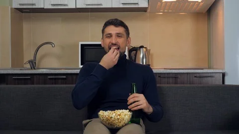 Man sitting on the couch, eats popcorn, drinking beer, smiles and watches TV Stock Footage 121841035