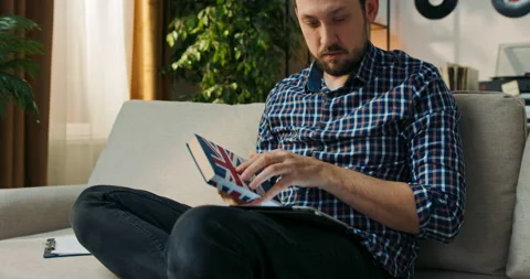 The man is sitting on couch at home doing homework for extra English classes Видео 200820089