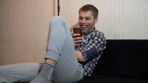 Man is sitting on the couch looking into the smartphone and laughing Stock-Footage 86160480