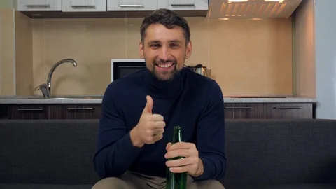 Man sitting on the couch, shows thumb up, takes beer, smiles and watches TV Stock Footage 121840766