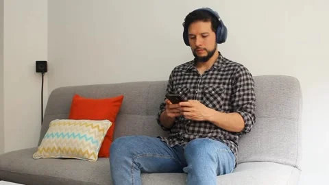 Man sitting on a couch using a wireless headset texting in his smartphone Stock Footage 340583407