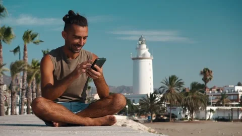 Man sitting cross-legged on wall using smartphone with lighthouse and beach.. Stock Footage 344059219