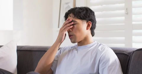 Man sitting on dark gray couch reading smartphone, rubbing forehead, snacking, Stock Footage 344386009