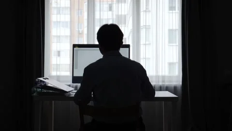 Man sitting at a desk in a dark room, working on a computer backlit by a window Stock Footage 345530813