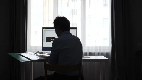 Man sitting at a desk in silhouette, working on a computer and holding a coffee Stock Footage 346250973