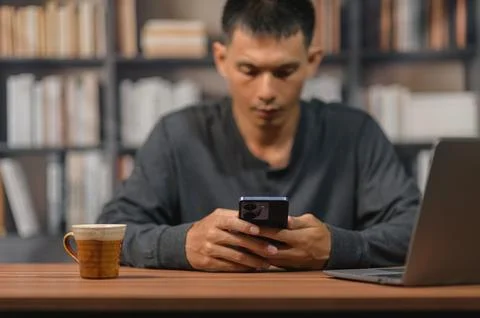 Man Sitting at Desk Using Smartphone in Office with Bookshelf Background Stock Photos