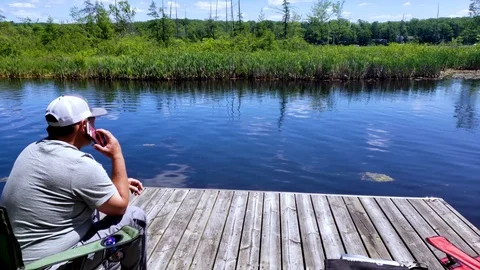 Man sitting on dock at the lake taking a phone call on iPhone or android Stock Footage 111370964