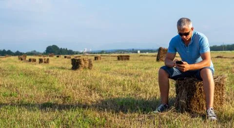 A man sitting in a field with a phone on a haystack. Stock Photos
