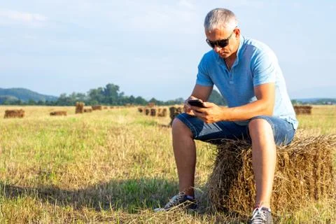 A man sitting in a field with a phone on a haystack. Stock Photos